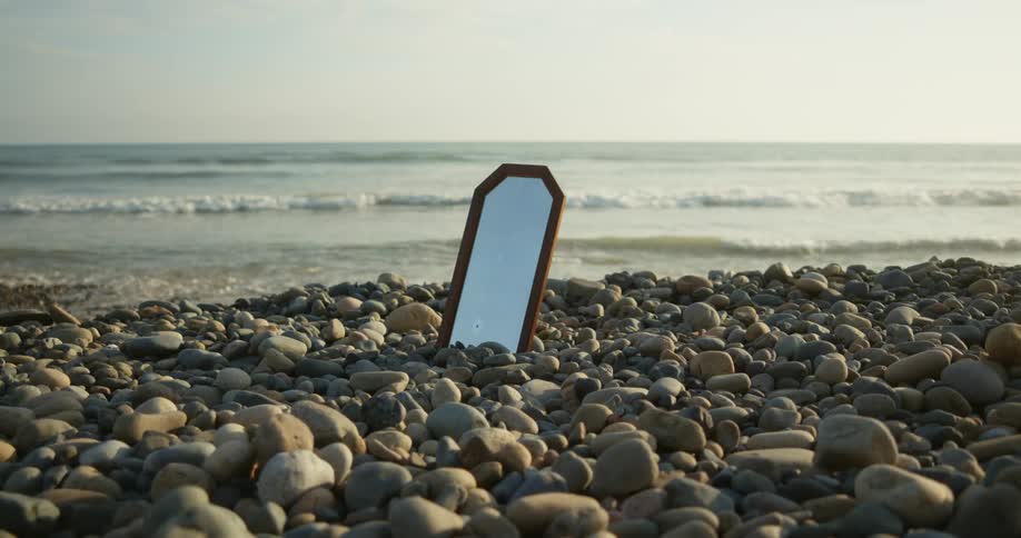 Waves roll onto pebble strewn beach as mirror set among pebbles reflects the sky