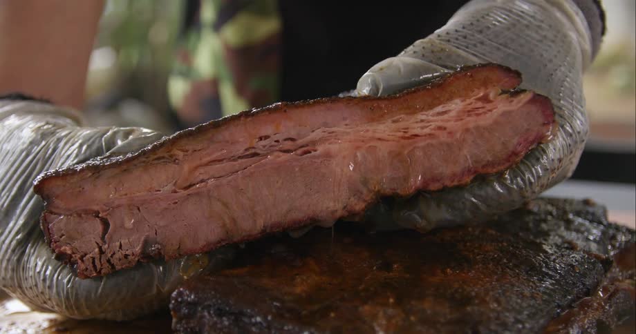 Gloved hands hold a barbecued slab of meat dripping with juices