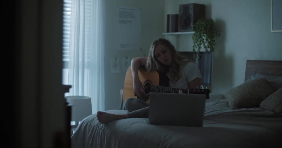 A teenage girl sits on her bed and plays the guitar