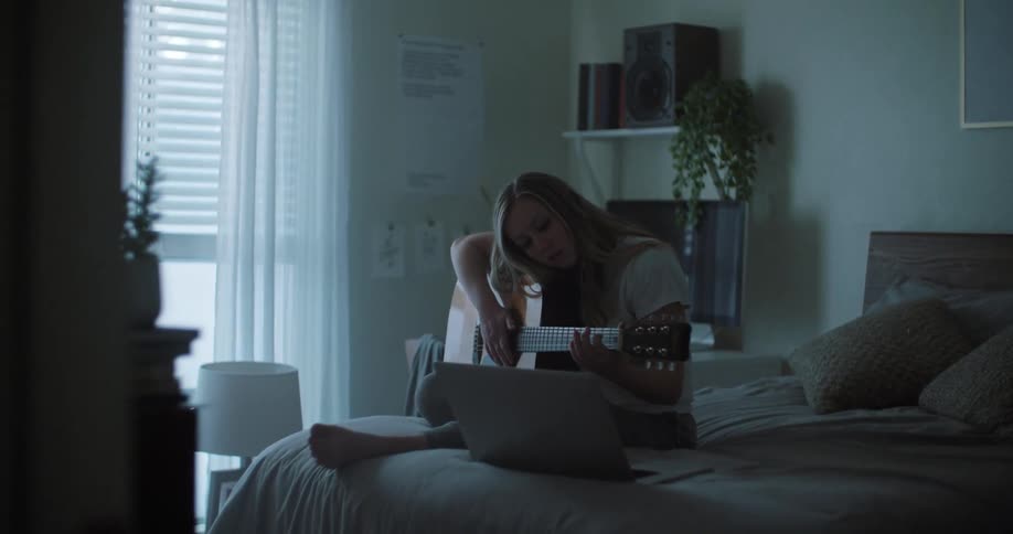 A teenage girl sits on her bed and plays the guitar