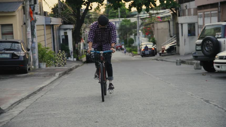Man pedals bicycle along road past parked vehicles and apartment buildings