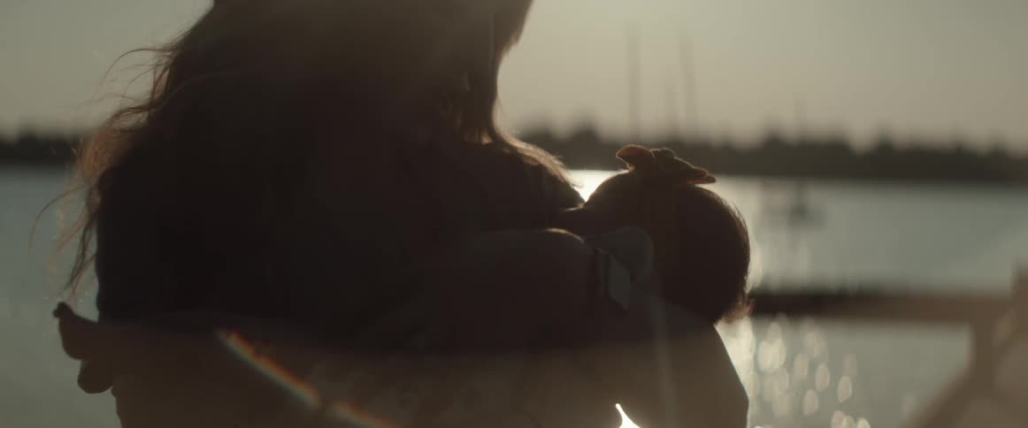 A mom cradling her daughter in her arms bends and kisses her while standing in the sunshine with water shimmering behind her