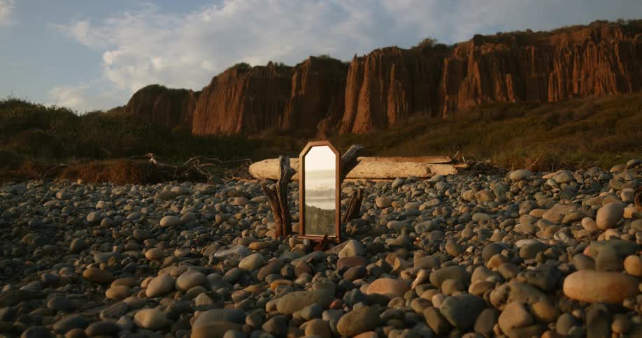 Mirror standing in field of pebbles reflects ocean surf with escarpment in background