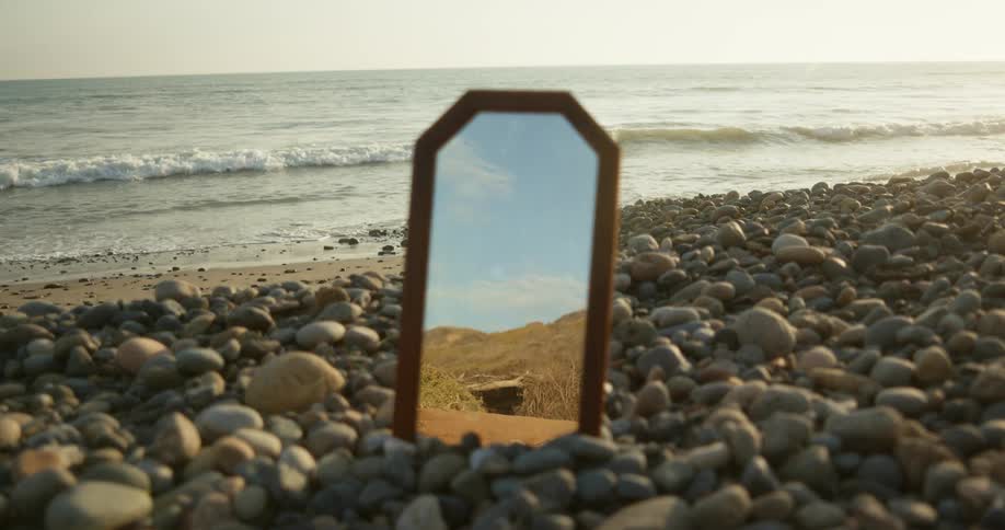 A mirror propped up in rocks on the shore shows a reflection of hills