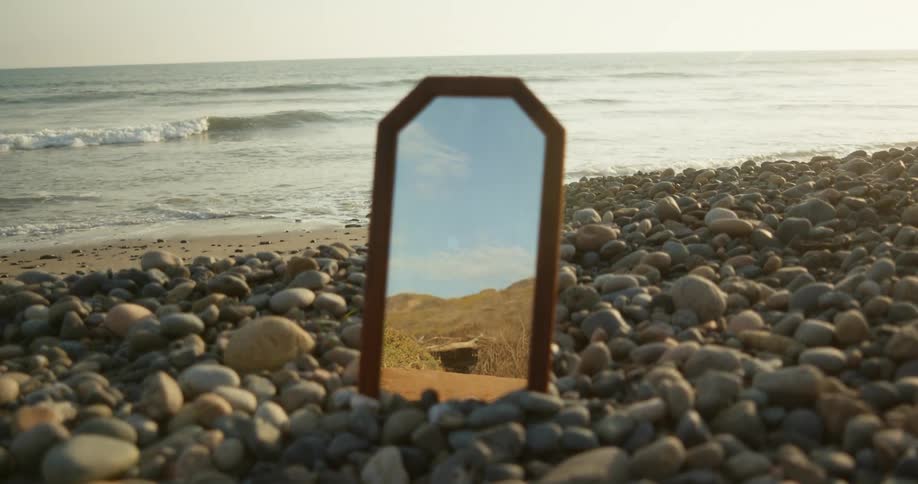 Mountains reflecting on a mirror standing on the rocks near the ocean at sunset