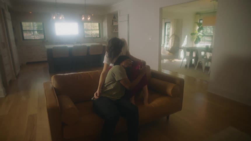 A man and a woman in a room on a couch hugging with a wall and doorway in the background