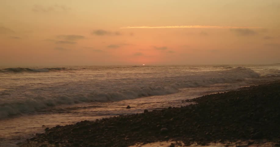 Sun sinks beneath the horizon in red sky as waves roll onto pebble strewn beach