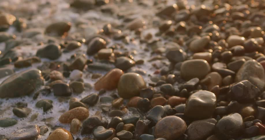 A small foamy wave washes up and over a pile of smooth rocks and pebbles on a beach