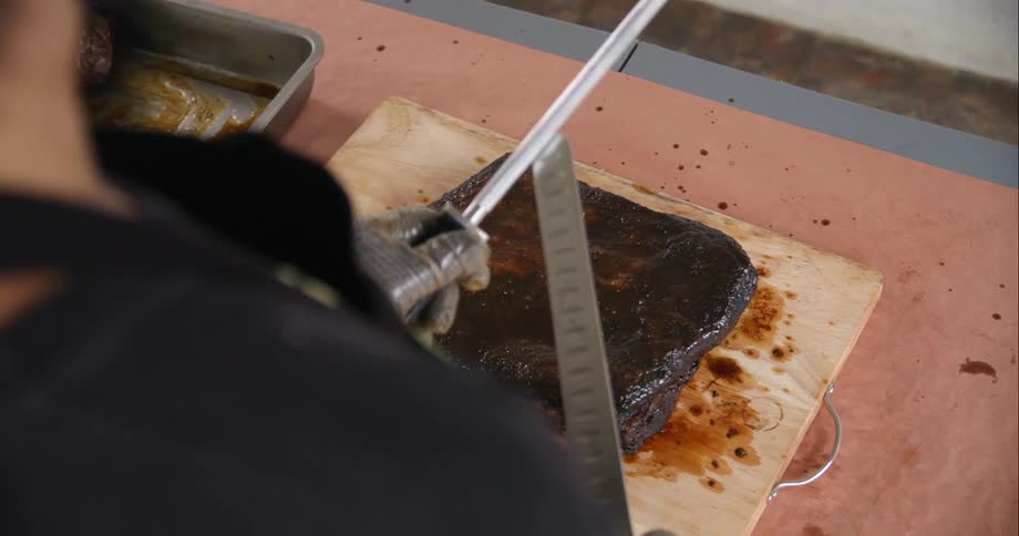 A man sharpens a carving knife above a slab of barbecued meat on a cutting board