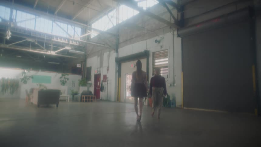 Two young women walk toward each other in a loft space and stand back to back while dancing