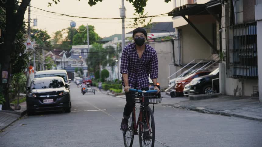 Man wearing face mask pedals bicycle along road past apartment buildings