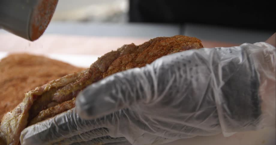 A person with gloves sprinkling seasoning on a piece of meat on a cutting board on a table