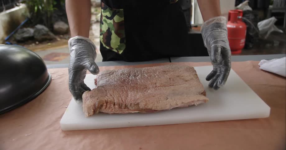 A man removes a cast iron lid covering a slab of brisket and flips the meat around
