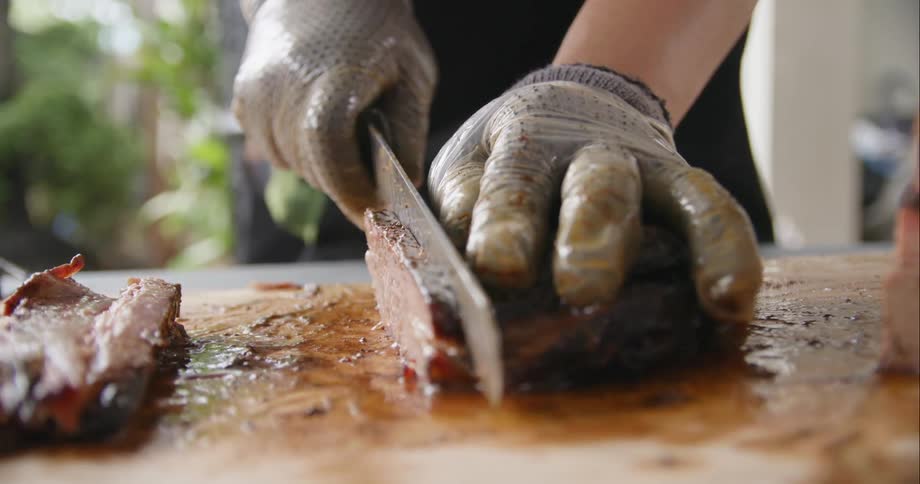 Person wearing gloves cuts beef on wooden board with a knife standing outdoors