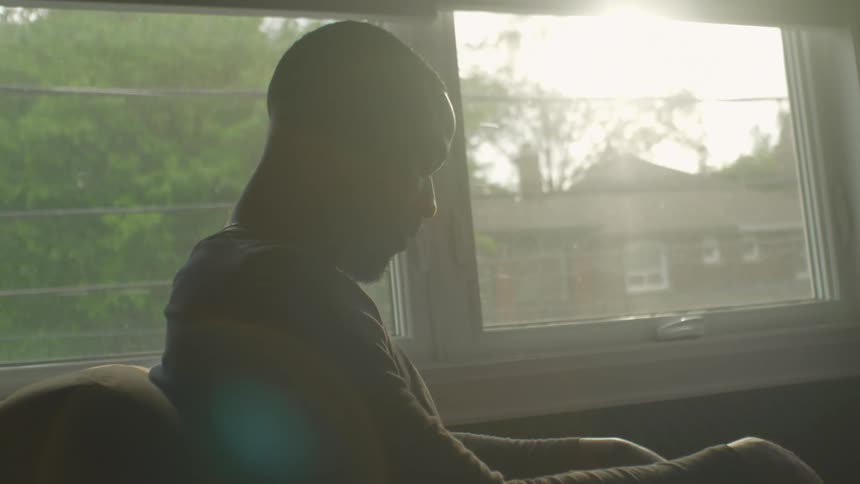 A man sits in a chair at home and looks down as he thinks
