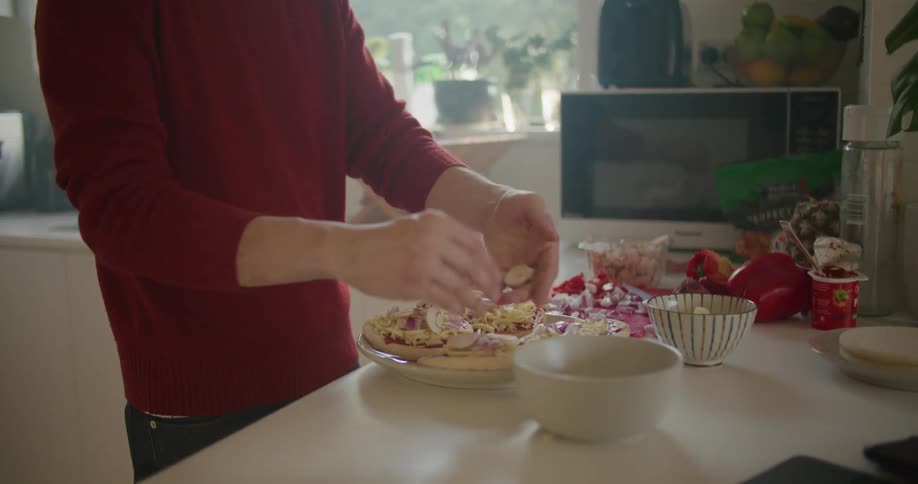 Man spreads cheese on mini pizzas standing in kitchen near microwave