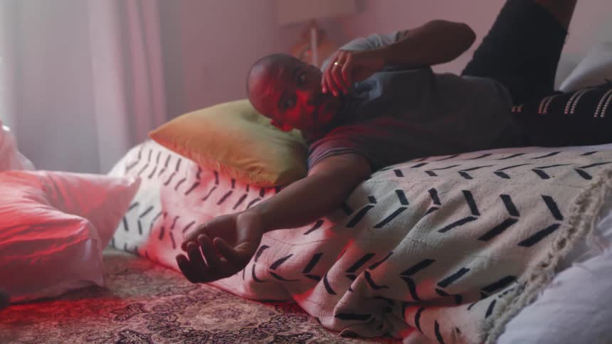 A man lies in bed at home and reaches over and pets his cat who then walks away and the man continues to watch his cat