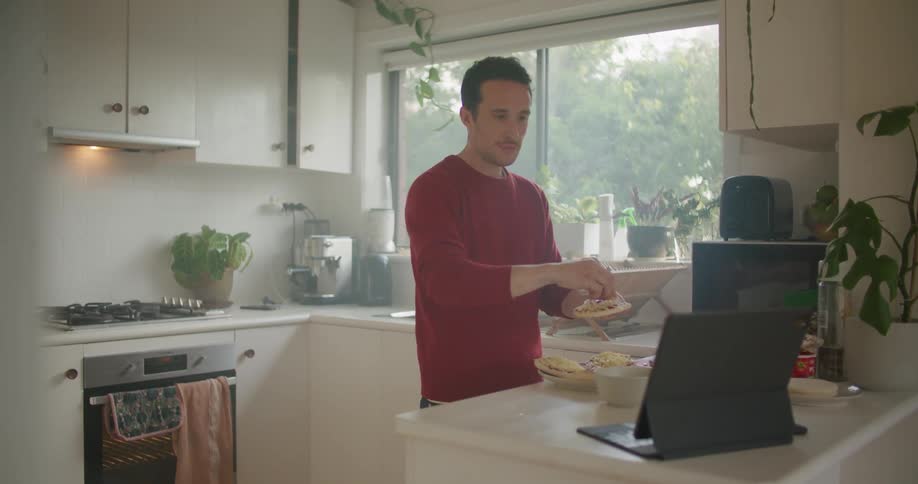 A man stands at a kitchen counter preparing food pausing to tap and look at a tablet