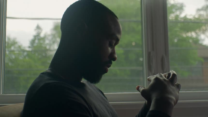 A man in a room sitting on a couch and praying with a window in the background