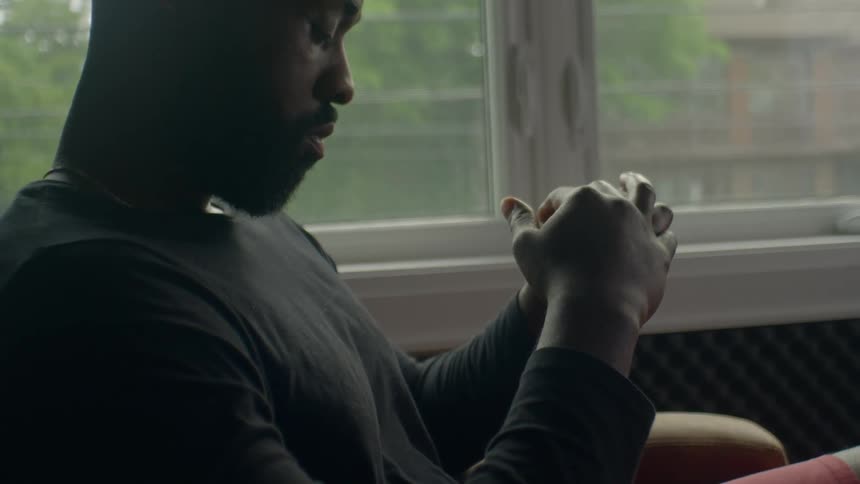 A man in a room sitting on a couch with a window and wall in the background