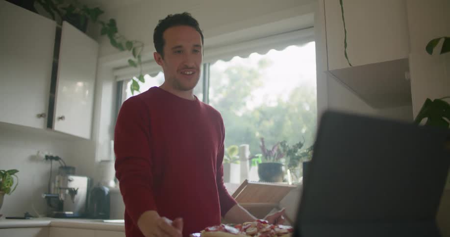 A man smiles and talks while he is preparing miniature pizzas in front of his laptop computer