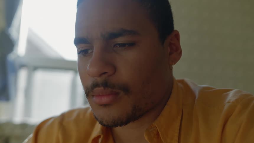 Man wearing yellow shirt sitting inside a room at home thinking