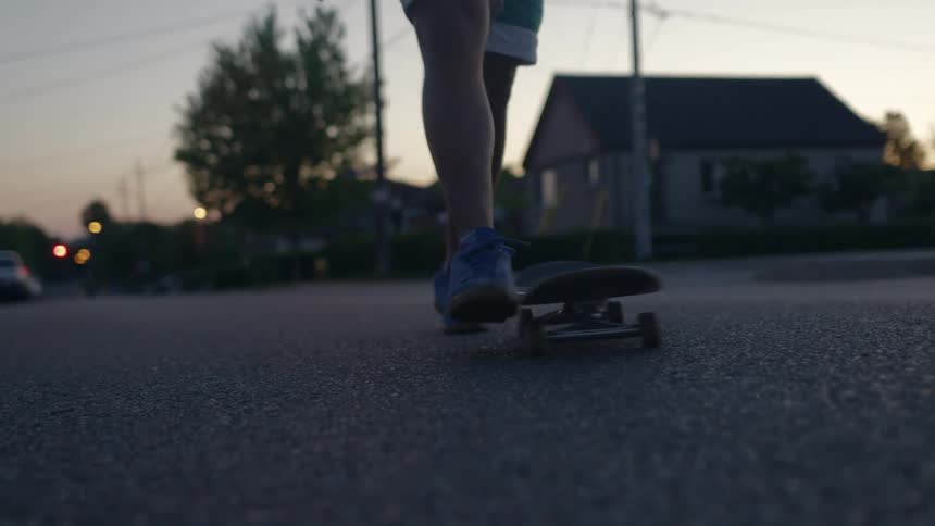 Man with white shorts orange shirts and blue sneakers skateboards on the road in the neighborhood at dusk