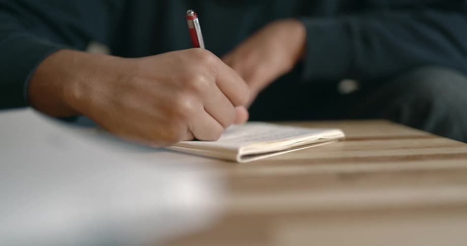 A man leans down on a coffee table as he writes in a small notebook