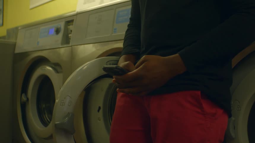 A man leans against a washing machine in a laundromat and types on his phone