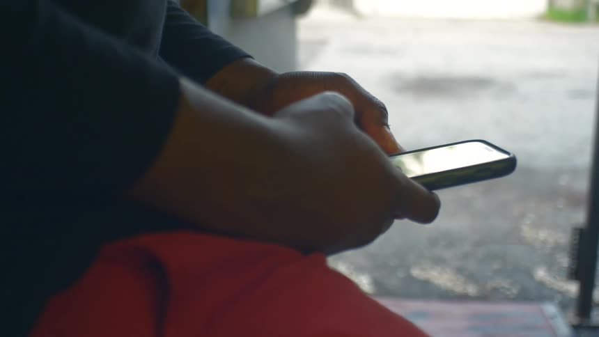 A man scrolls on his phone using his thumb while leaning against a wall outside