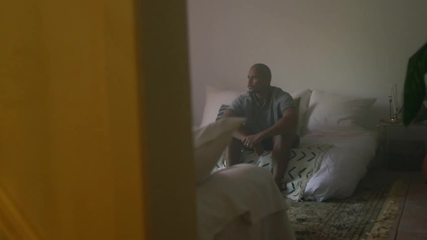 A man sits on his bed at home and looks towards the window
