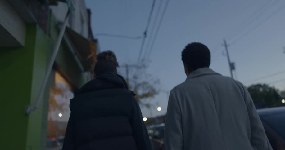 Two women are walking along a city sidewalk at dusk with cars passing in the background