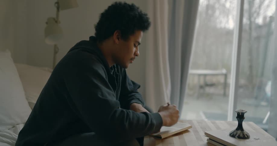 A man pauses to think about what to write while sitting on the couch with a small notebook