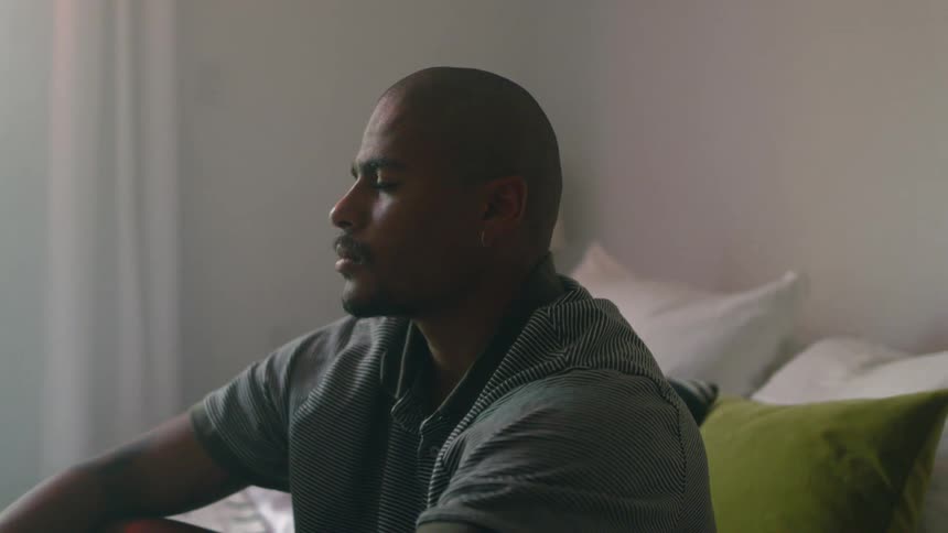 Man with mustache and shaved head sits on bed pondering