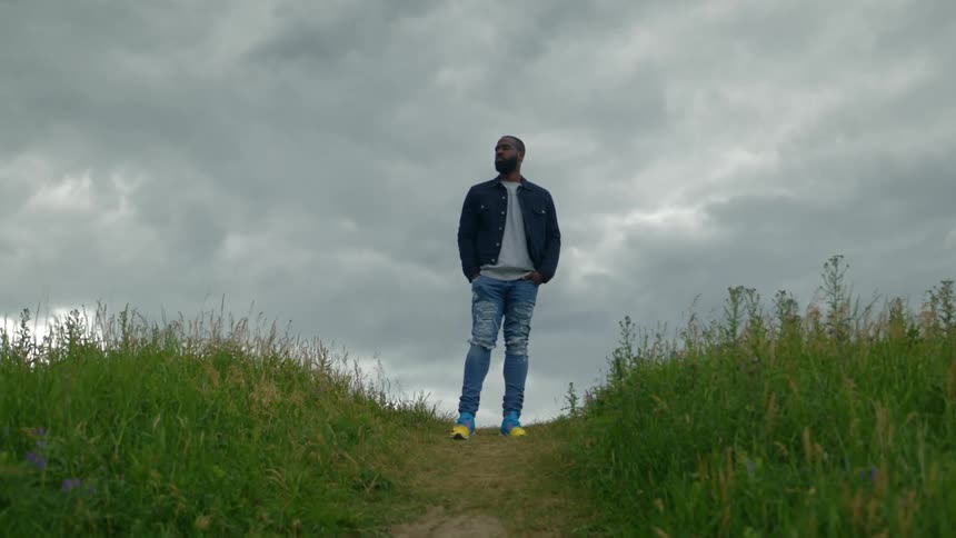 Man with a beard jacket and jeans stands on dirt road on hill on a cloudy day