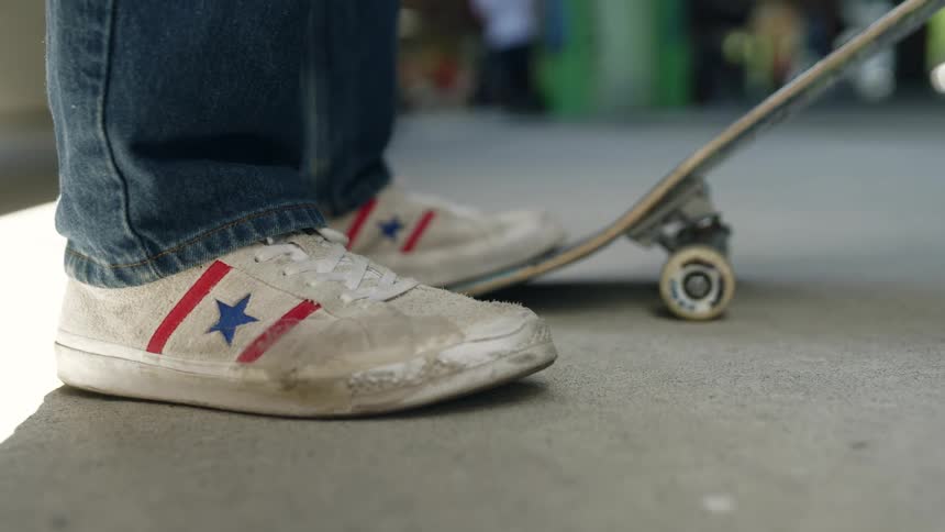 Person stands with one foot on end of skateboard as people mill around in the background
