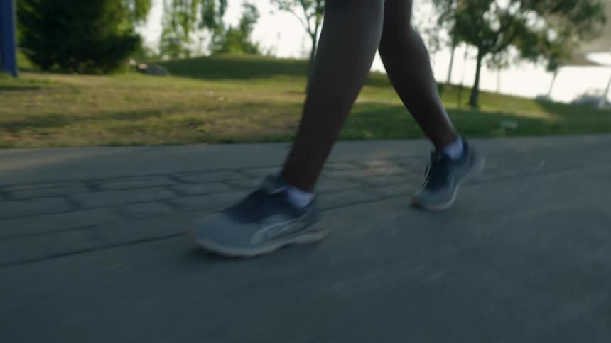 A person wearing running shoes walks briskly along a paved pathway next to a park