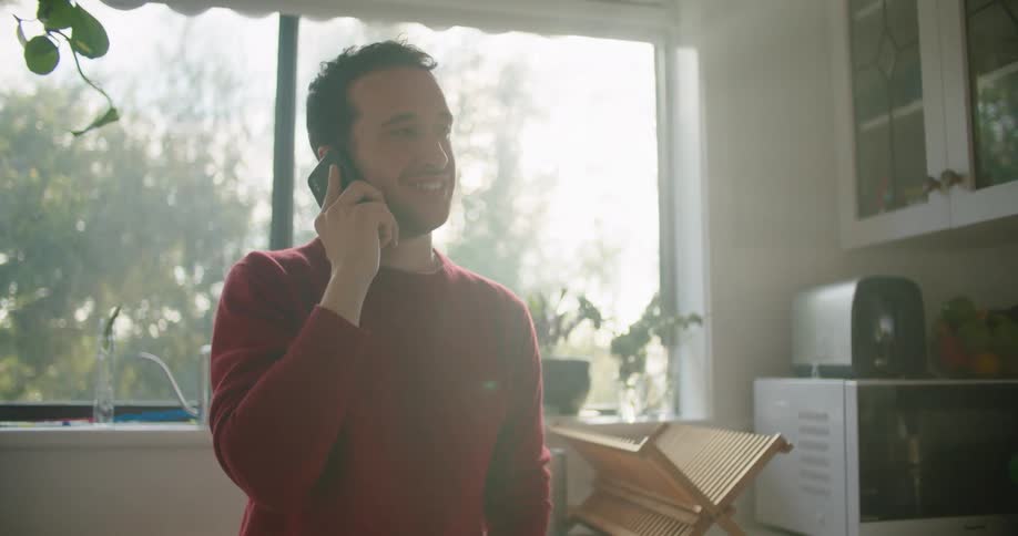 A man stands in the kitchen talking on the phone smiling and laughing
