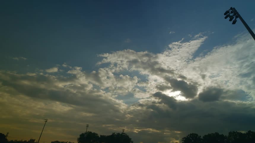 The sun is obscured by a layer of clouds above a park with towering poles with light fixtures