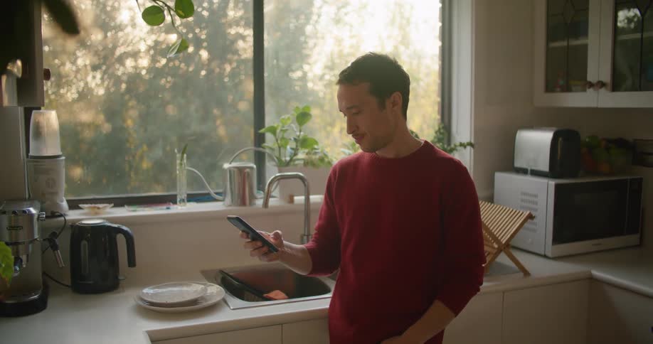Man stands in kitchen near window showing a tree as he scrolls on mobile phone