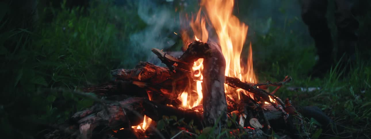 A man with a hat and glasses stoking a fire with a knife with trees the sky and clouds in the background