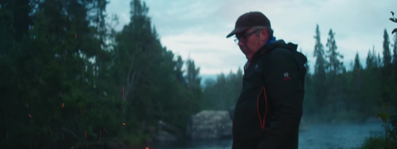 A man stands next to a river and turns to a fire on the shore