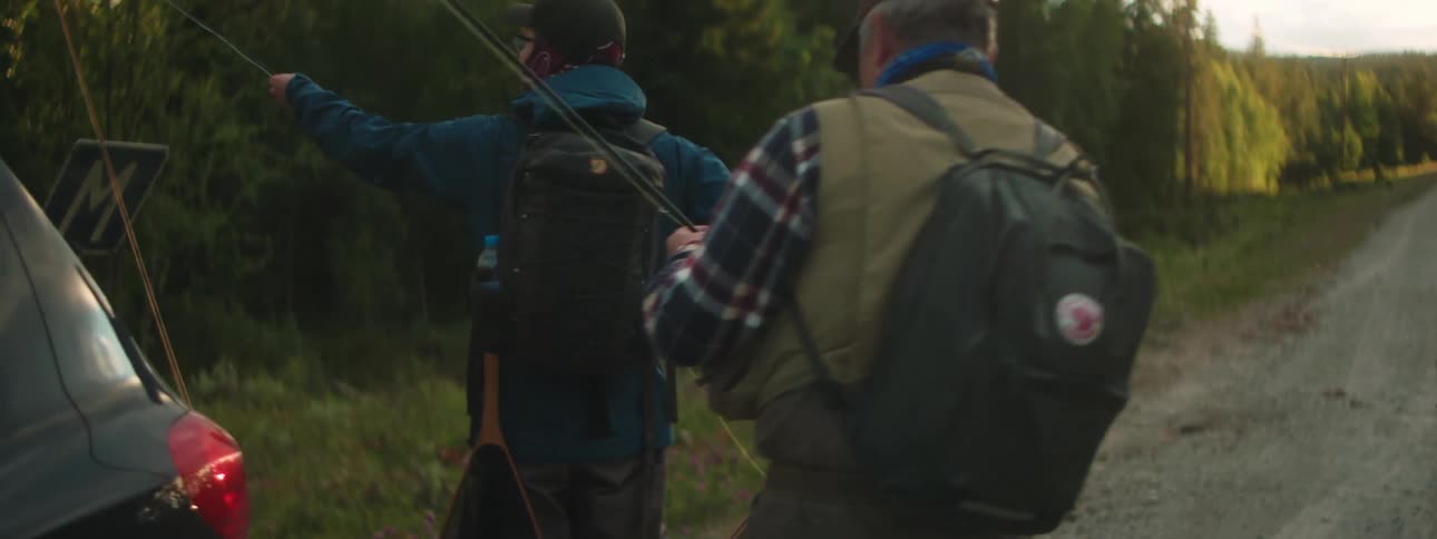 Two fishermen walk around back of car parked on side of dirt road through forest