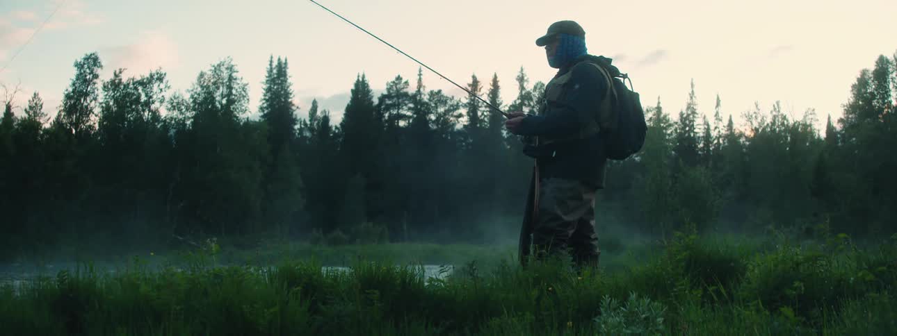 Man wearing backpack and baseball cap stands on bank of river fishing with forest in background