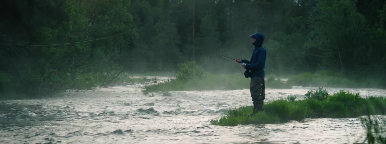 Angler casts fishing line standing near river on misty day as sun shines between trees in woods