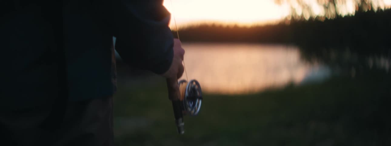 A person holding a fishing rod with a lake trees and a cabin in the background