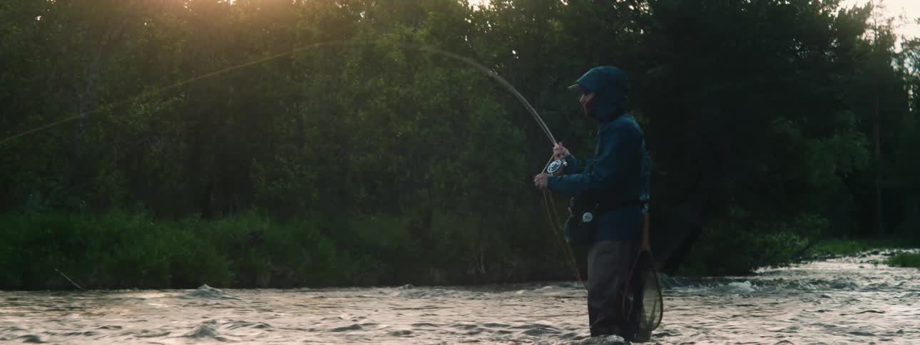 A man standing in a shallow river is cast a fishing line in the early morning
