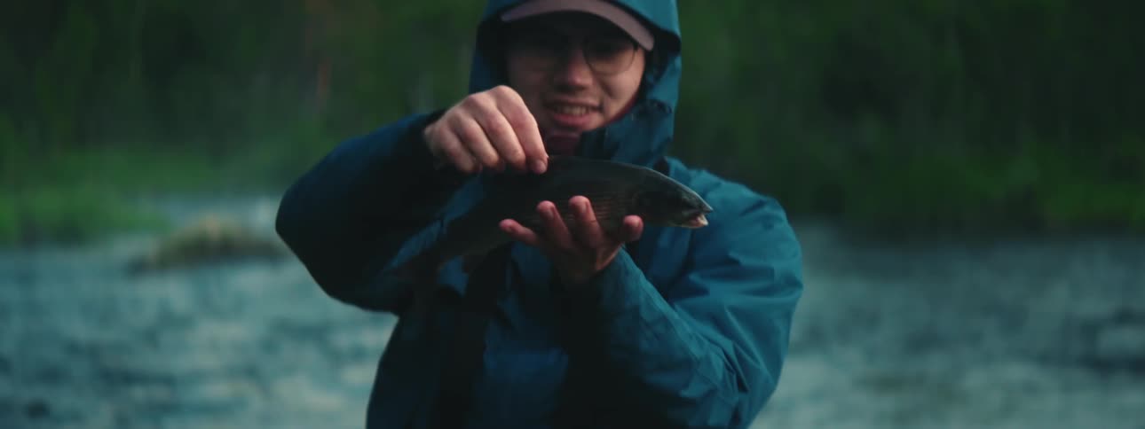A man is standing by a river in a forest happily presenting a fish that he caught in the early morning