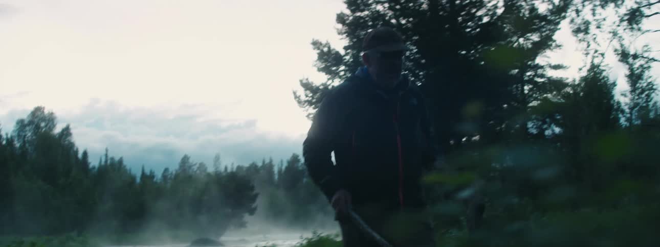 A man with a hat walking with branches towards a fire with trees the sky and clouds in the background