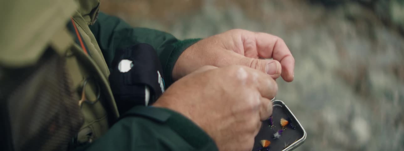 A fisherman opens a small case to select a fishing fly